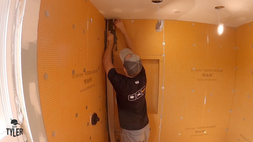 man using drywall knives to apply Kerdi wrap to corner seam of waterproofing board